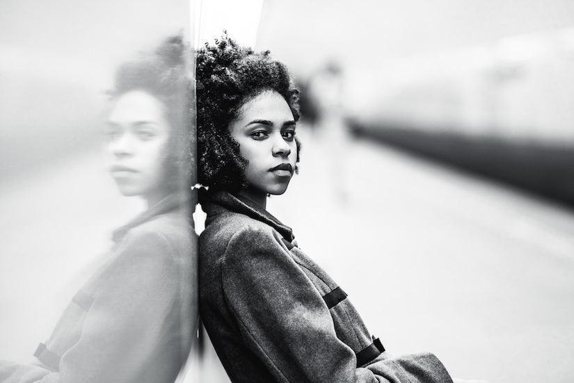 A black and white portrait of an African American female in a coat leaning against a mirror wall; she has a sombre reflective look on her face. Image for an article: 