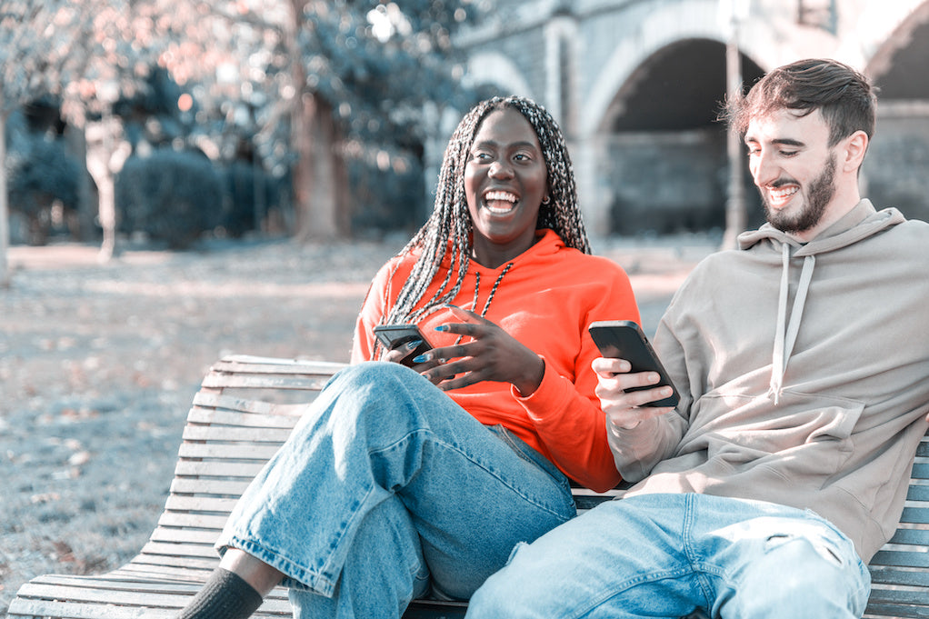 Happy multi ethnic young couple looking mobile phone at park sitting on bench laughing about funny responses to How are you?