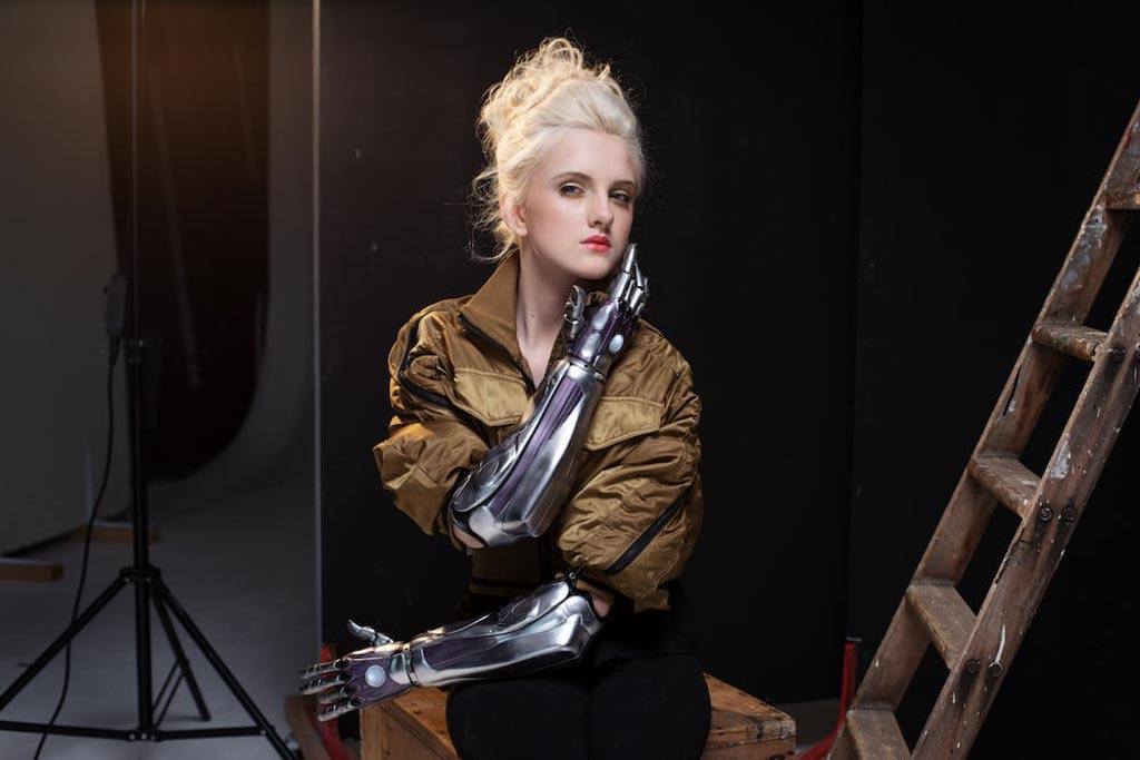 Photo of Tilly Lockey, bionic arm girl, on the set of a photoshoot. Her body faces forward as she looks at the camera. She is wearing an army green oversized bomber jacket and black trousers.