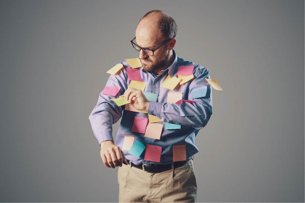 Labels belong on bottles not people: a man in a long-sleeve shirt is covered in post-it-labels and is removing them, one by one.