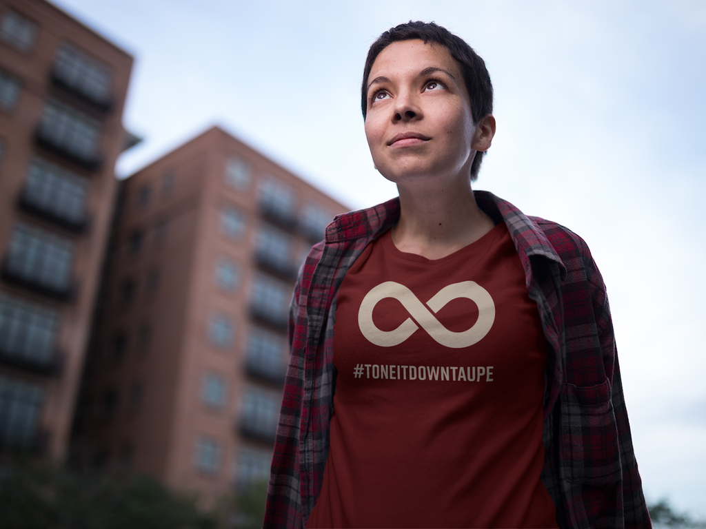 This is a photo of a person wearing an oxblood black Tone It Down Taupe shirt standing outside. In the middle top one-third of the tee is a taupe colored infinity symbol. Beneath the symbol is the hashtag word printed in bold upper case letters: #ToneItDo