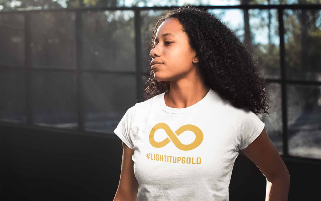 A Black person with an afro wearing a white Light It Up Gold t-shirt. The tee has the word #LightItUpGold printed in large upper case letters beneath a solid line infinity symbol. The symbol and text are colored gold.