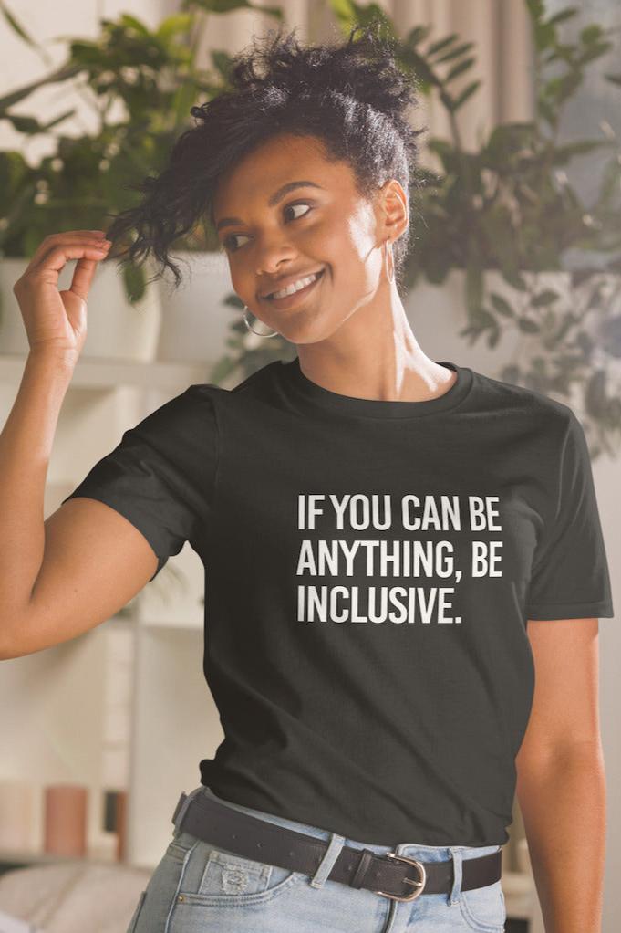 This is the photo of a black Be Inclusive Basic Tee. On the front of the t-shirt, the phrase, 'If you can be anything, be inclusive,' is printed in white capital letters. The tee is worn by a Black woman.