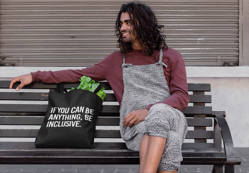 Smiling man sitting on a bench with a black tote bag that reads ‘If You Can Be Anything, Be Inclusive.’ Shop diversity bags, inclusion totes, and backpacks.