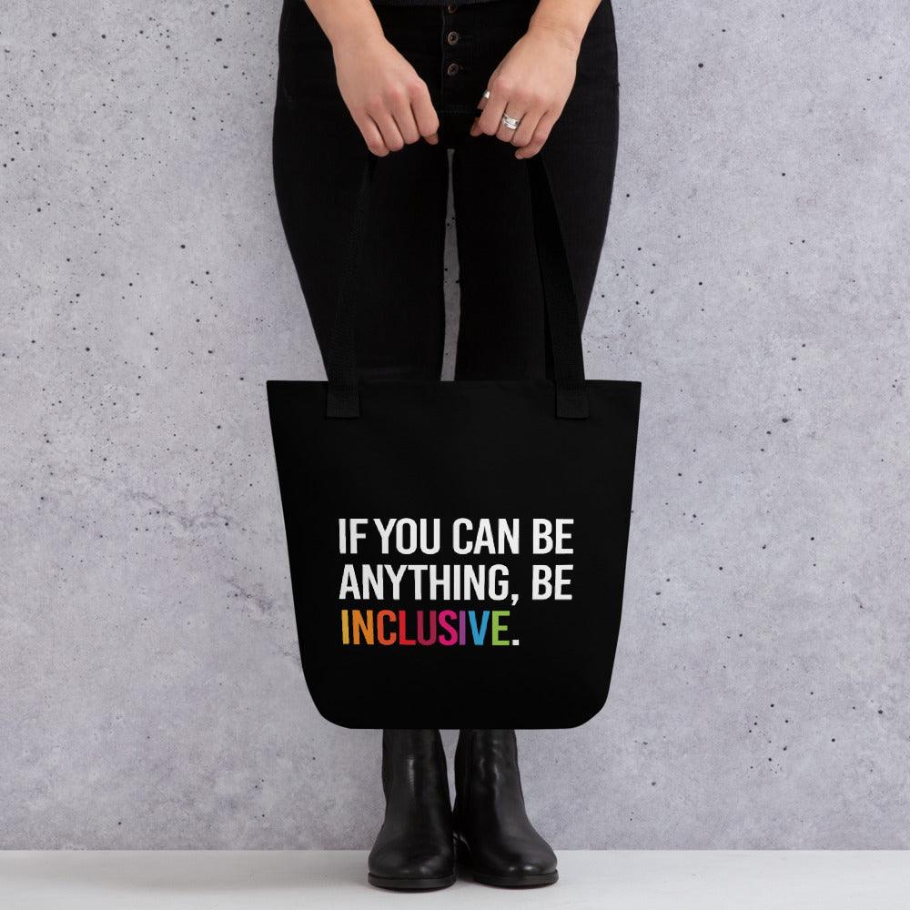 Inclusive Tote Bag. The black tote has the phrase, 'If you can be anything, Be Inclusive,' printed in white. The word inclusive is in the colors of the rainbow. The inclusive tote is being held by a person in front of their knees.